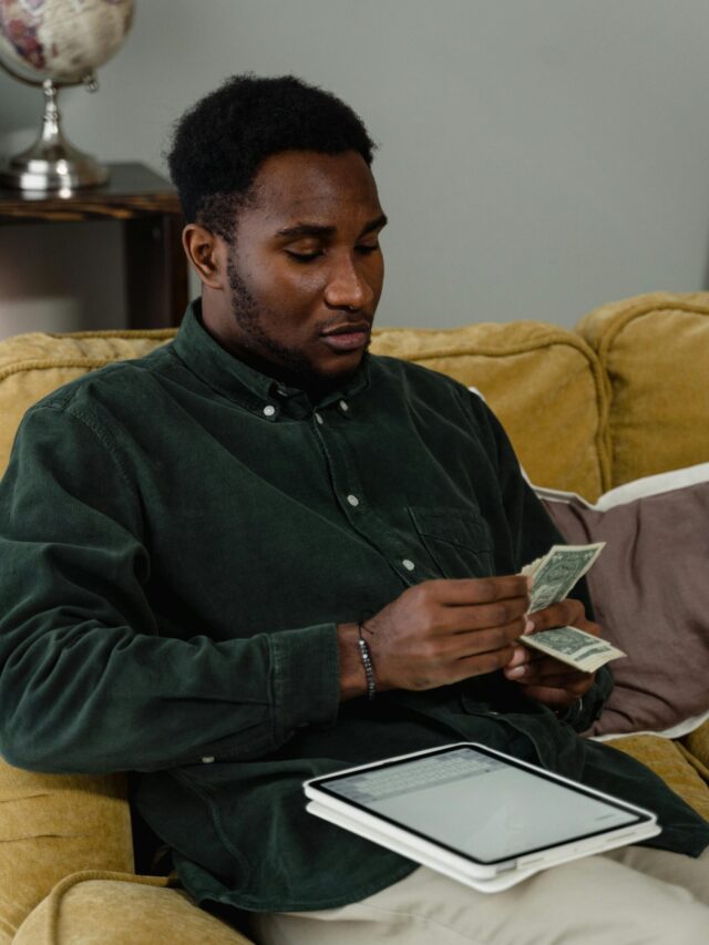 A businessman counting cash while seated on a sofa with a tablet at hand, reflecting on financial management.