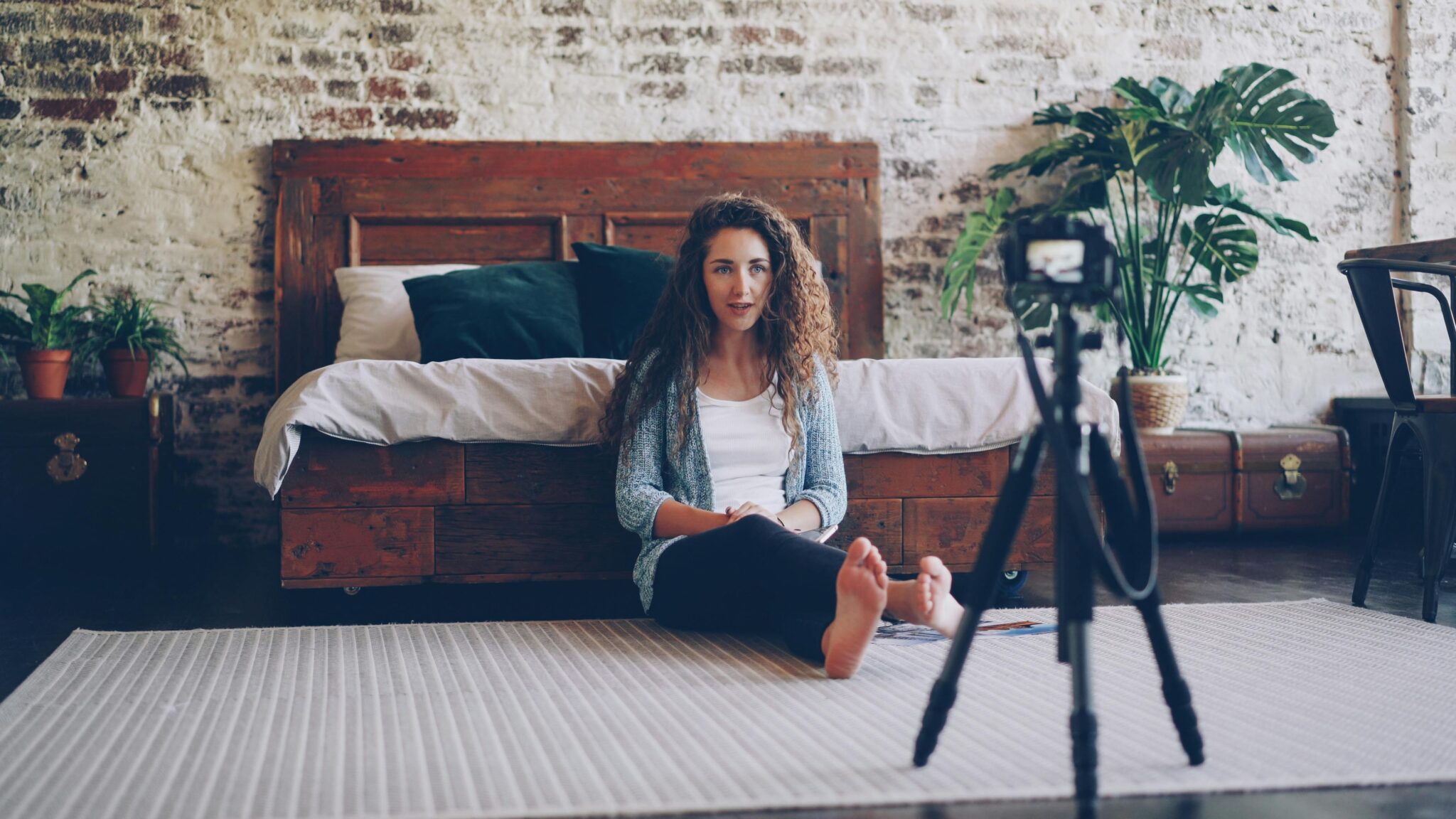 Woman seated in bedroom with camera, creating content in relaxed environment.